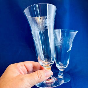 May include: Two clear, etched champagne flutes with floral and leaf designs. The flutes have a slender, elongated shape with a flared top and a round base. One flute is held by a hand, and the background is a solid blue.