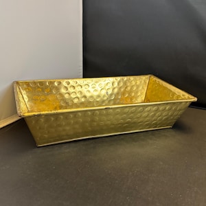 May include: A rectangular, gold-colored metal planter with a hammered texture. The planter has a slightly trapezoidal shape, with a flat base and angled sides. The interior is empty, and the planter is set against a dark background.