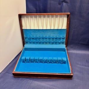 May include: Open wooden silverware storage box with a blue velvet interior. The box has a dark wood frame, a cream-colored fabric lining the lid, and compartments for utensils.
