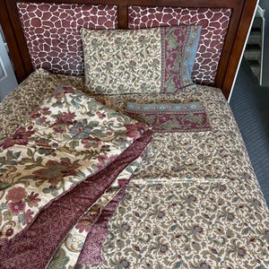 May include: A bed with a dark wood frame and a headboard featuring a giraffe print. The bedding includes a floral patterned comforter, pillowcases, and a decorative pillow with a floral and geometric design. The color palette includes cream, burgundy, and teal.