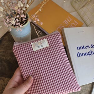 Könnte beinhalten: Eine rot-weiß karierte Reißverschlusstasche mit einem cremefarbenen Etikett mit der Aufschrift "FREDAMA". Die Tasche wird in einer Hand gehalten, mit einem Buch und einer Vase mit Blumen im Hintergrund. Die Tasche wird wahrscheinlich zur Aufbewahrung kleiner Gegenstände verwendet.