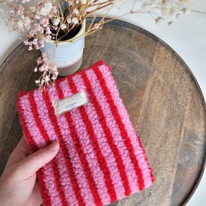 Peut inclure: Pochette rectangulaire à rayures roses et rouges, duveteuse, avec une étiquette « FRIDAVA ». La pochette est posée sur un plateau en bois, avec un petit vase de fleurs séchées en arrière-plan.
