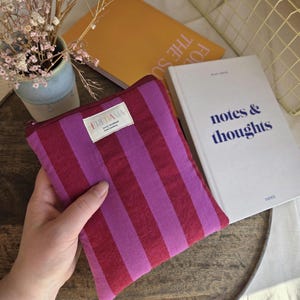 Op de afbeelding: Een rechthoekig etui met ritssluiting en verticale strepen in rood- en paars tinten. De etui heeft een klein wit label met de tekst "FREDAMA". De etui wordt in een hand gehouden, met een boek en andere items op de achtergrond.
