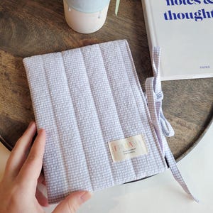 Peut inclure: Une couverture de livre en vichy lavande et blanc avec une étiquette blanche sur laquelle est inscrit "FRIDAMA" et "Small Handmade Bookbinding" en rose. La couverture est ornée d'un ruban blanc.