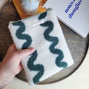 Könnte beinhalten: Eine weiße, strukturierte Tasche mit einem teal Reißverschluss und teal, wellenförmigen Mustern. Die Tasche hat ein kleines Etikett mit der Aufschrift "FRIDA". Im Hintergrund ist ein Buch mit den Worten "notes & thoughts" zu sehen.