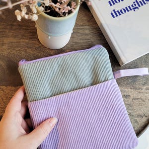 Könnte beinhalten: Eine hellgrüne und lavendelfarbene Reißverschlusstasche aus Cordstoff. Die Tasche ist in der Hälfte gefaltet und wird in einer Hand gehalten. Die Tasche befindet sich auf einer Holzoberfläche.