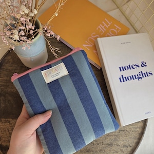Könnte beinhalten: Ein blau-grau gestreifter Reißverschlussbeutel mit rosa Reißverschluss, beschriftet mit "FRIDAMA". Der Beutel wird in der Nähe eines weißen Buches mit "notes & thoughts" auf dem Cover und eines gelben Buches mit Text gehalten.