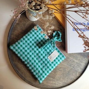 Könnte beinhalten: Eine grün-weiß karierte Tasche mit passender Schleife. An der Tasche ist ein kleines rechteckiges Etikett befestigt. Die Tasche liegt auf einem Holztablett mit einem Buch und Trockenblumen.