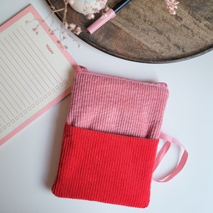 Könnte beinhalten: Zwei Cord-Reißverschlusstaschen, eine hellrosa und eine rote, sind auf einer weißen Oberfläche gestapelt. Ein rosa Notizblock mit dem Wort "TODAY" ist sichtbar, zusammen mit einem Holztablett und einem rosa Stift. Die Taschen haben eine gerippte Textur.