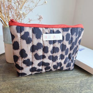 Könnte beinhalten: Eine gesteppte Kosmetiktasche mit einem Leopardenmuster in Schwarz und Beige. Die Tasche hat einen roten Reißverschluss und eine rote Einfassung. Auf dem Etikett steht "FRIDA".