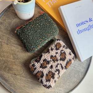 Könnte beinhalten: Zwei kleine Reißverschlusstaschen. Eine ist strukturiert grün mit orangefarbenen Flecken, die andere hat einen tan- und braunen Leopardenmuster. Beide liegen auf einem Holztablett mit einem kleinen Keramiktopf und Büchern im Hintergrund.