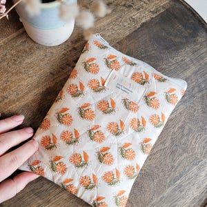 Könnte beinhalten: Eine kleine, gesteppte Reißverschlusstasche mit einem Blumendruck in Orange- und Grüntönen. Die Tasche hat einen weißen Hintergrund und ein Etikett mit der Aufschrift "Every Handmade from Handbags".