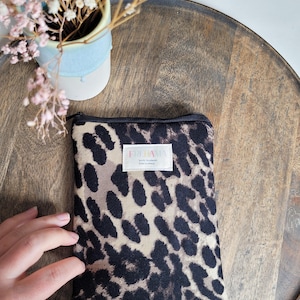 Könnte beinhalten: Kleine, rechteckige Tasche mit schwarzem Reißverschluss und Leopardenmuster in Schwarz und Beige. Das weiße Etikett trägt den Text "FREDAMIA". Die Tasche liegt auf einer Holzoberfläche.