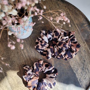 Può includere: Tre elastici per capelli con stampa leopardata nei toni del marrone, nero e crema. Gli elastici per capelli in tessuto morbido sono disposti su un vassoio di legno. Un piccolo vaso con fiori rosa è sullo sfondo.