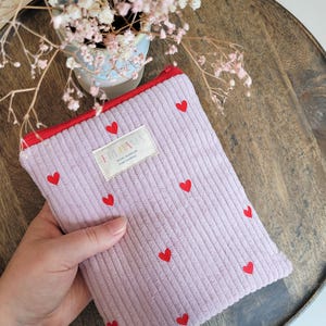 Könnte beinhalten: Ein lavendelfarbenes Cord-Etui mit roten Herzakzenten. Das Etui hat einen roten Reißverschluss und ein Etikett mit der Aufschrift "FRIDANA". Das Etui liegt auf einem Holztablett mit einer kleinen Vase mit Trockenblumen.
