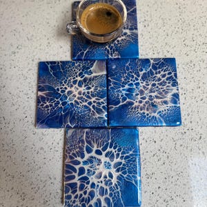 May include: Set of four blue and white coasters with a swirling, abstract pattern. The coasters are made of resin and have a glossy finish.