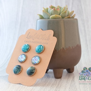 May include: A set of three pairs of stud earrings featuring a variety of designs. The earrings are displayed on a brown card with the text "Leaf&Chick" and "LeafAndChick.com".