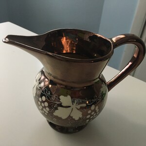 May include: A copper-colored pitcher with a rounded body and a spout. The pitcher features a handle and is decorated with a white grape vine design. The pitcher is likely used for serving beverages.