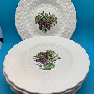 May include: A stack of eight white ceramic dinner plates with a raised embossed design. The plates feature a grapevine design with purple grapes and green leaves in the center.