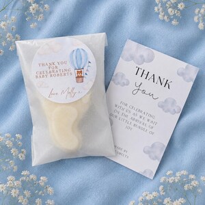 May include: A translucent bag containing a cream-coloured foot-shaped wax melt with a round sticker featuring a hot air balloon and a bear, reading Thank You For Celebrating Baby Roberts Love Molly x, next to a card reading Thank You For Celebrating With Us As We Wait On The Arrival Of Our Little Bundle Of Joy.