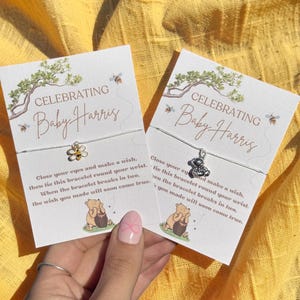 May include: Two cards with the text "CELEBRATING Baby Harris" and a bee and bear charm bracelet. The cards have a watercolor tree and bee design. The text on the card reads "Close your eyes and make a wish..."