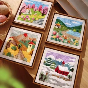 May include: Four framed embroidered landscape scenes. One depicts a campsite with pink trees, another a house by a lake, a third a windmill, and the last a red barn in a snowy scene. Each artwork is in a wooden frame.