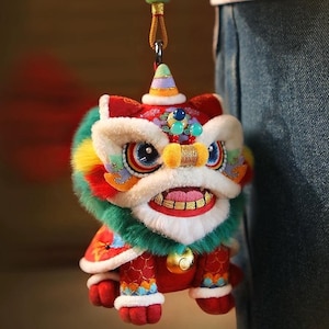 Könnte beinhalten: Ein farbenfrohes Löwentanz-Ornament aus Plüsch mit detailliertem Design. Der Löwe hat rote, weiße, grüne, gelbe und blaue Akzente, eine goldene Glocke und eine Aufhängeschlaufe. Eine traditionelle chinesische Neujahrsdekoration.