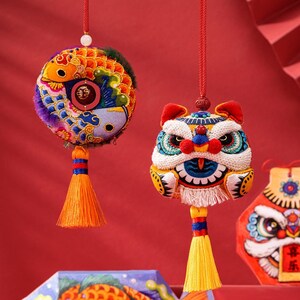 以下が含まれることがあります： 2つのカラフルな中国のお正月の飾り、1つはライオンの頭の形、もう1つは2匹の魚がいる円の形状をしています。両方の飾りは複雑な刺繍が施されており、赤い紐に黄色の房がついた状態で吊り下げられています。