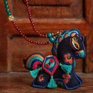 Peut inclure: Pendentif cheval brodé noir avec des accents colorés, orange, bleu et vert. Le pendentif est attaché à un collier de perles rouges avec un fermoir vert et doré. Le cheval présente un design détaillé avec des pompons.