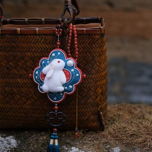 May include: A white felt rabbit charm with blue and red embroidery, hanging from a brown woven basket with a red beaded necklace. The charm features a pearl and a blue tassel.