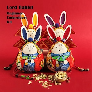 May include: Four colorful embroidered rabbit-shaped pouches with white fur, red, blue, and yellow accents. The pouches are arranged in a row on a red background. The text "Lord Rabbit Beginner Embroidery Kit" is visible at the top of the image.