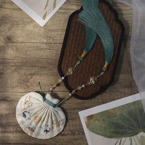 Może przedstawiać: Kremowa torebka w kształcie wachlarza z haftowanymi kwiatowymi wzorami, połączona z łańcuszkiem z koralików i turkusowymi frędzlami. Torebka leży na drewnianej powierzchni z elementami dekoracyjnymi.