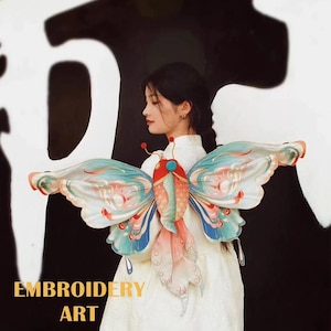 May include: A woman wearing a white dress with a large, colorful butterfly embroidered on the back. The butterfly is made of fabric and has a fish-like body with wings that are decorated with intricate patterns. The butterfly is a beautiful example of embroidery art.