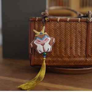 Op de afbeelding: Een kleine, geborduurde stoffen hanger met een aapgezichtontwerp. De hanger is bevestigd aan een lange, gele kwast en hangt aan een geweven bamboemand.