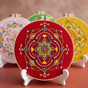 Può includere: Una collezione di cerchi ricamati colorati. Il cerchio centrale è rosso con un design simmetrico dettagliato in giallo, blu e bianco. Altri cerchi presentano motivi floreali e geometrici in rosa, verde e giallo, tutti su supporti bianchi.