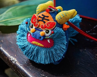 May include: A blue and red embroidered lion dance head with yellow ears and a blue fringe. The head is attached to a red string.