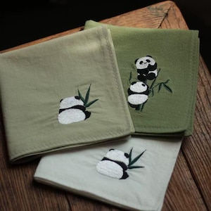 Pode incluir: Três lenços quadrados em tons de verde e branco, bordados com desenhos de pandas. Os pandas são pretos e brancos, com folhas de bambu. Os lenços estão dobrados e dispostos sobre uma superfície de madeira.