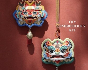 Può includere: Due ornamenti ricamati colorati a forma di cane-leone con dettagli intricati, appesi su uno sfondo marrone-rossastro. Ogni ornamento presenta una campana e un nappa. Il testo "DIY EMBROIDERY KIT" è visibile.