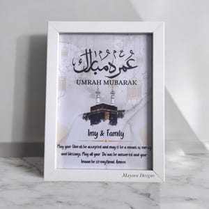 May include: White framed print with a white background and gold accents. The print features Arabic calligraphy that reads "Umrah Mubarak" and a watercolor illustration of the Kaaba. The print also includes the text "Imy & Family" and a prayer for a successful Umrah pilgrimage.
