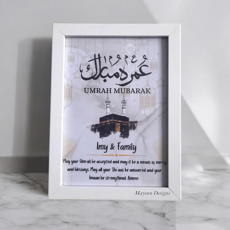 May include: White framed print with a white background and gold accents. The print features Arabic calligraphy that reads "Umrah Mubarak" and a watercolor illustration of the Kaaba. The print also includes the text "Imy & Family" and a prayer for a successful Umrah pilgrimage.