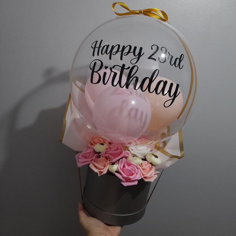 Pu&ograve; includere: Un bouquet di palloncini trasparente con un palloncino rosa e pesca all'interno. Il palloncino ha una scritta nera che dice "Happy 23rd Birthday". Il bouquet &egrave; in una scatola grigia con un arrangiamento di fiori rosa e bianchi.