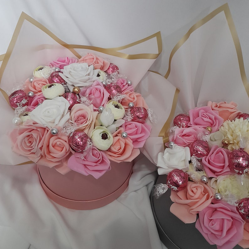 Pu&ograve; includere: Due scatole regalo rosa piene di rose rosa e bianche e caramelle al cioccolato. Le scatole sono avvolte in carta velina dorata e bianca.