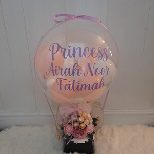 Pu&ograve; includere: Un palloncino trasparente a forma di mongolfiera con un palloncino rosa, bianco e rosa chiaro all'interno. Il palloncino ha un nastro viola legato intorno alla parte superiore e le parole "Princess Airah Noor Fatimah" scritte su di esso. Il palloncino &egrave; seduto su una scatola nera con un fiocco rosa e bianco e un bouquet di fiori rosa e cioccolato.