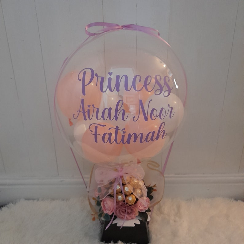 Pu&ograve; includere: Un palloncino trasparente a forma di mongolfiera con un palloncino rosa, bianco e rosa chiaro all'interno. Il palloncino ha un nastro viola legato intorno alla parte superiore e le parole "Princess Airah Noor Fatimah" scritte su di esso. Il palloncino &egrave; seduto su una scatola nera con un fiocco rosa e bianco e un bouquet di fiori rosa e cioccolato.