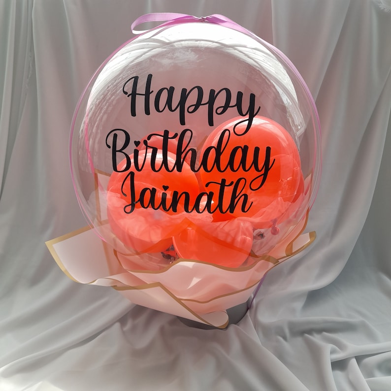 Pu&ograve; includere: Un bouquet di palloncini trasparente con il testo "Happy Birthday Jainath" stampato su di esso. Il palloncino &egrave; riempito con palloncini arancioni pi&ugrave; piccoli.