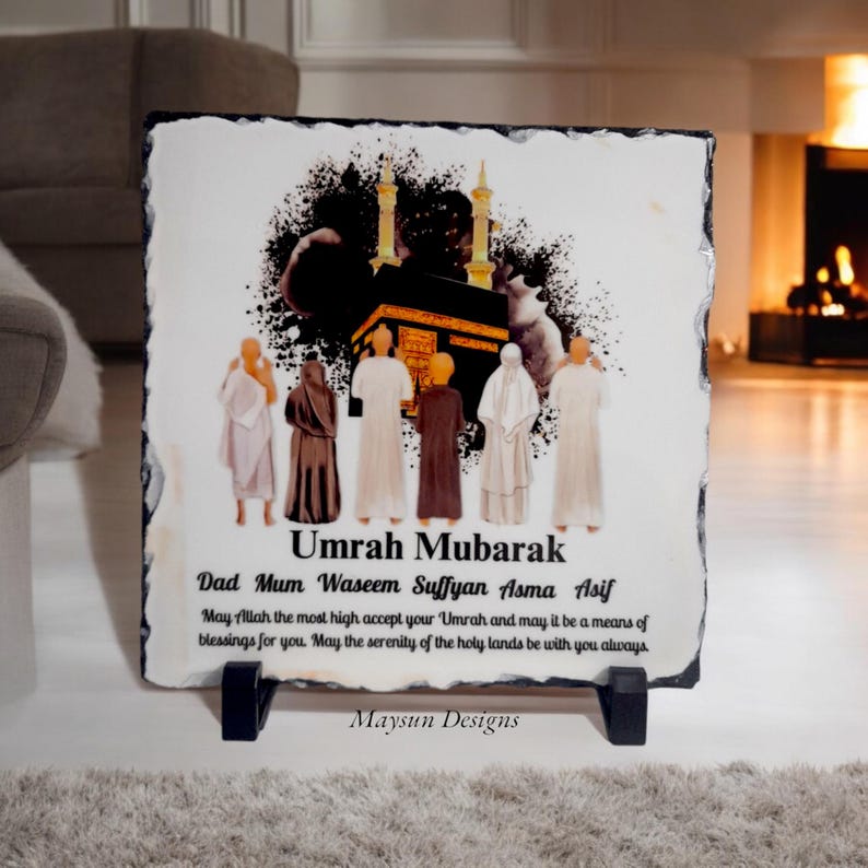 May include: A white slate plaque with a black and gold illustration of the Kaaba and people praying. The text "Umrah Mubarak" is written in black at the top of the plaque. Below the illustration, the text "Dad Mum Waseem Suffyan Asma Asif" is written in black. Below that, a message in black text reads "May Allah the most high accept your 'Umrah and may it be a means of blessings for you. May the serenity of the holy lands be with you always." The text "Maysun Designs" is written in black at the bottom of the plaque.