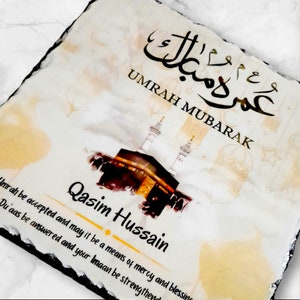May include: A white slate plaque with a watercolor illustration of the Kaaba in Mecca, Saudi Arabia. The plaque has the Arabic text "Umrah Mubarak" and the English text "Umrah be accepted and may it be a means of mercy and blessing. Du'aas be answered and your Imaan be strengthened." The plaque also has the name "Qasim Hussain" written in gold lettering.