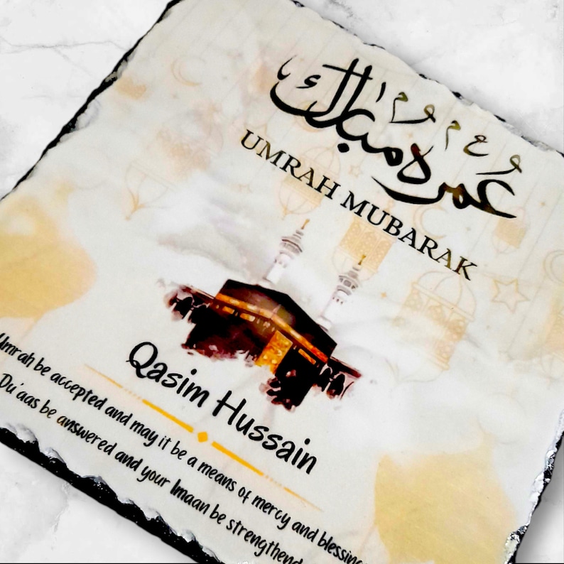 May include: A white slate plaque with a watercolor illustration of the Kaaba in Mecca, Saudi Arabia. The plaque has the Arabic text "Umrah Mubarak" and the English text "Umrah be accepted and may it be a means of mercy and blessing. Du'aas be answered and your Imaan be strengthened." The plaque also has the name "Qasim Hussain" written in gold lettering.