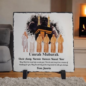 May include: A black slate plaque with a colourful illustration of the Kaaba, a mosque in Mecca, Saudi Arabia. The plaque has the text "Umrah Mubarak" and a message wishing someone a blessed Umrah pilgrimage. The text also includes the names "Narmin Rameen Nawal Noor" and "Javeria".