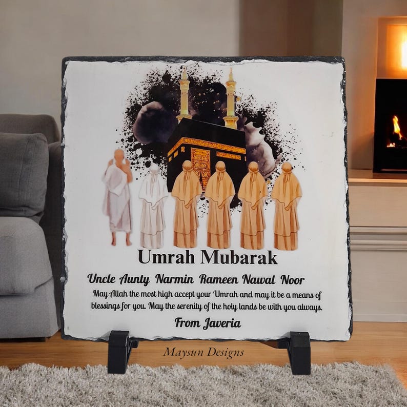 May include: A black slate plaque with a colourful illustration of the Kaaba, a mosque in Mecca, Saudi Arabia. The plaque has the text "Umrah Mubarak" and a message wishing someone a blessed Umrah pilgrimage. The text also includes the names "Narmin Rameen Nawal Noor" and "Javeria".